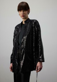 Čierny flitrovaný kabát s stojacím golierom, voľným strihom a dlhými rukávmi. Jemne lesklá textúra zvyšuje vizuálny efekt látky.