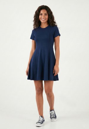 Femeie zâmbitoare, purtând o rochie cu mâneci scurte, de culoare albastru închis, și adidași albaștri închiși cu șireturi albe, stând pe un fundal alb simplu.