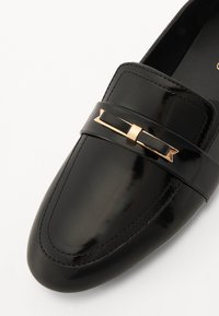 Melnas patentētas ādas loafers ar plakanu dizainu, kas ietver zelta toņu dekoratīvā apdare pāri vampam un smalkas šuvju detaļas.