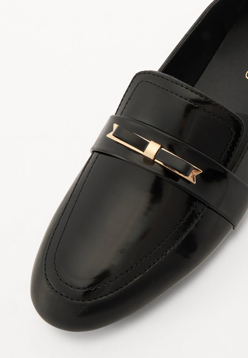 Schwarze Lackleder-Loafer mit flachem Design, ausgestattet mit einer goldfarbenen Dekoration auf dem Vorderblatt und dezenten Nähdetails.
