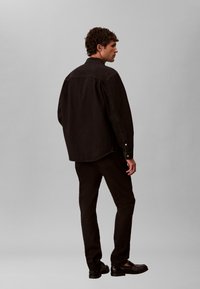 Άνδρας με σγουρά μαλλιά που φοράει μαύρο μακρυμάνικο πουκάμισο, μαύρο παντελόνι και μαύρα παπούτσια, στέκεται μπροστά σε απλό ανοιχτό γκρι φόντο.