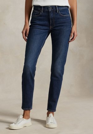 Osoba nosi priliehavé tmavomodré džínsy a biele tenisky na šnurovanie, stojaca na neutrálom pozadí.