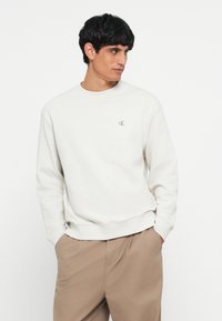 Lys beige genser med rund hals og ribbede mansjetter. Har en liten grå 'CK'-logo på venstre bryst. Myk tekstur.