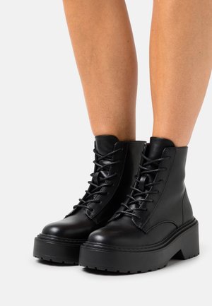 Botines de cuero negro hasta el tobillo con puntera redondeada, cordones en la parte delantera y suela de plataforma gruesa. Textura suave con detalles de costura robusta.