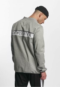 Mann iført en grå jakke med "IKKE KJØR MED OSS" på ryggen og en svart lue, ser nedover mot en ensfarget bakgrunn.