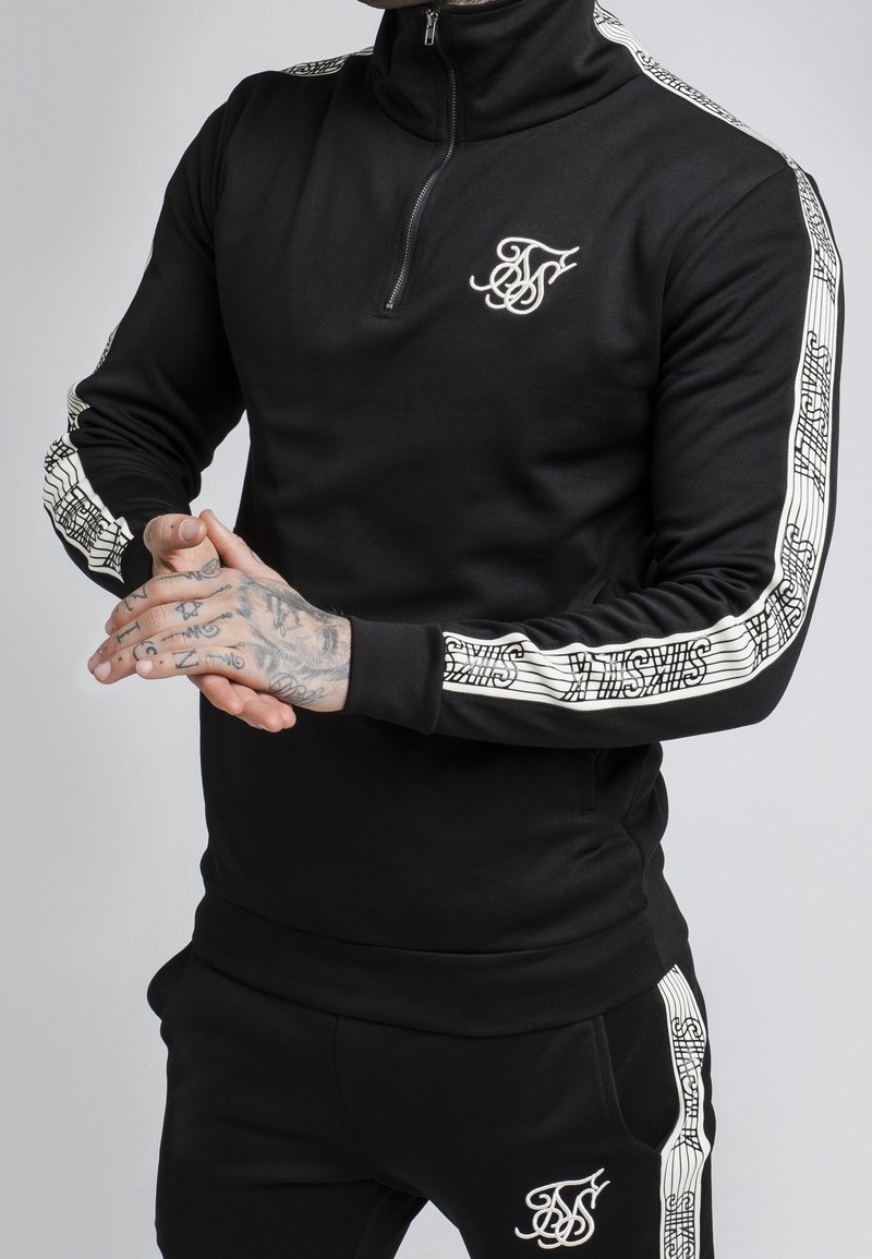 Black zip-up sports top with white logo and patterned accents on the sleeves, paired with matching black pants. Smooth, stretchy fabric.