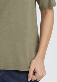 Olivzaļas krāsas adīta t-krekls ar īsām piedurknēm, apaļu izgriezumu un taisnu apakšu, valkāts kopā ar džinsa biksēm. Redzama gluda tekstūra.