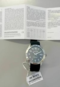 Orologio argento con quadrante grigio, marcatori delle ore bianchi e cinturino in gomma nera. Include un'etichetta e un manuale utente sullo sfondo.