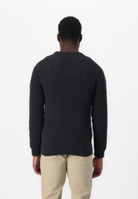 Ciemny sweter dziany o fakturowanej powierzchni, z okrągłym dekoltem oraz ściągaczami przy rękawach i dolnym brzegu, zestawiony z beżowymi spodniami. Widok od tyłu.