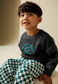 Šedá mikina s tyrkysovým nápisem "BIG HAPPY SMILE" a odpovídajícím úsměvem. Bílé a zelené kostkované kalhoty s měkkou texturou.