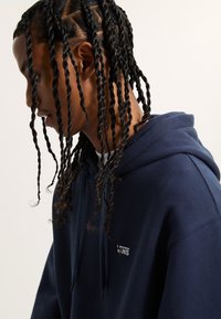 Felpa con cappuccio blu navy dotata di tasca a marsupio, polsini a coste e un discreto dettaglio del logo ricamato. Capelli lunghi e intrecciati visibili, con volto leggermente inclinato verso il basso.