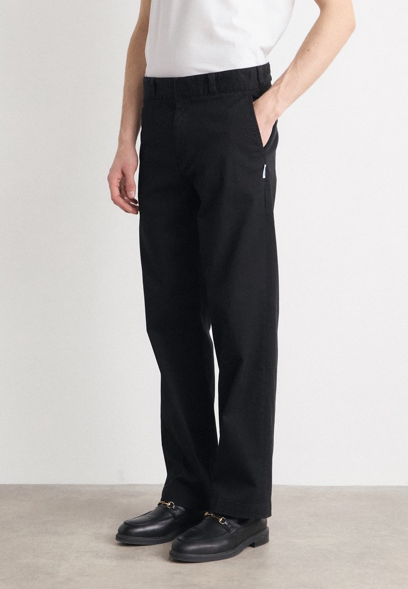 Homme portant un pantalon droit noir avec poches latérales et des mocassins en cuir noir, debout devant un fond clair uni.