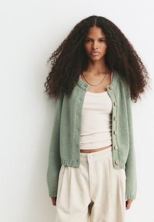 Jeune femme avec de longs cheveux bouclés portant un cardigan vert clair sur un débardeur beige et un pantalon beige taille haute, se tenant devant un fond blanc.