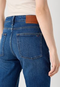 Nærbilde av en kvinnes midje som har på seg blå jeans med en synlig lærmerkelapp som sier "TOMMY JEANS" på bakbuksen.