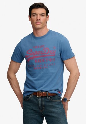 Man draagt een blauw SuperDry T-shirt met rode tekst, donkere spijkerbroek, een bruine leren riem en een horloge met een blauwe wijzerplaat om zijn linkerpols.