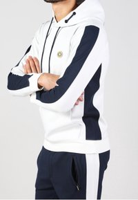 Felpa con cappuccio bianca e pannelli laterali blu navy. Presenta un cappuccio regolabile con cordino e un logo circolare sul petto. Tessuto morbido, vestibilità casual.