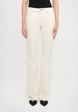 Donna che indossa jeans a gamba dritta bianchi a vita alta con chiusura frontale e scarpe nere con punta appuntita su uno sfondo neutro.