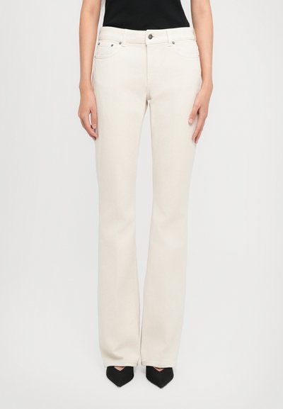 Kvinde iført højtskårne off-white straight-leg jeans med forreste knap og sorte spidse sko mod en ensfarvet baggrund.
