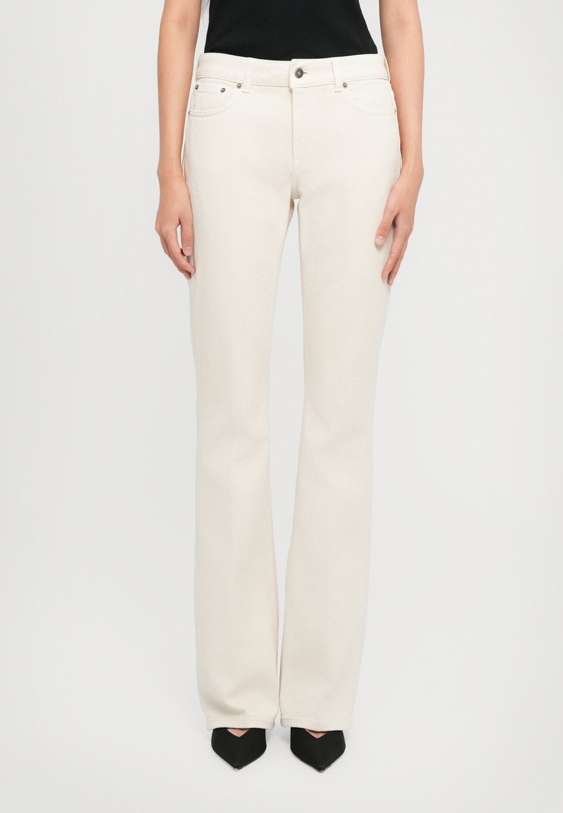 Mujer que lleva puestos unos pantalones rectos de tiro alto de color blanco roto, con botón frontal, y zapatos negros de punta against un fondo liso.