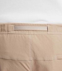 Beige byxor med en mjuk textur, med en vävd midja och kontrasterande vit söm. Två bakfickor är synliga.