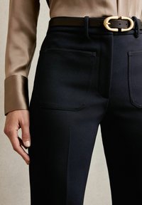 Schwarze maßgeschneiderte Hose mit glatter Textur und zwei Gesäßtaschen, kombiniert mit einem beigen Seidenhemd und einem schwarzen Gürtel mit goldener Schnalle.