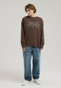 Brązowa koszula z długim rękawem z grafiką, luźne niebieskie dżinsy i białe sneakersy, stojąca na neutralnym tle.