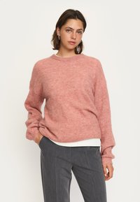 Rosa ulltröja med avslappnad passform, rund halsringning och långa ärmar. Buren över en vit skjorta och ihop med grå byxor.
