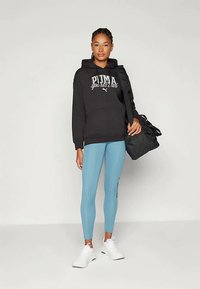 Sweatshirt noir à capuche avec impression "PUMA sports lab", associé à des leggings bleu clair et des baskets blanches. Portant un sac de sport noir.