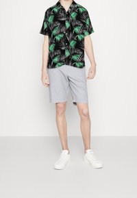 Kortärmad svart skjorta med mönster av gröna palmblad, tillsammans med ljusgrå shorts. Modellen bär vita sneakers.