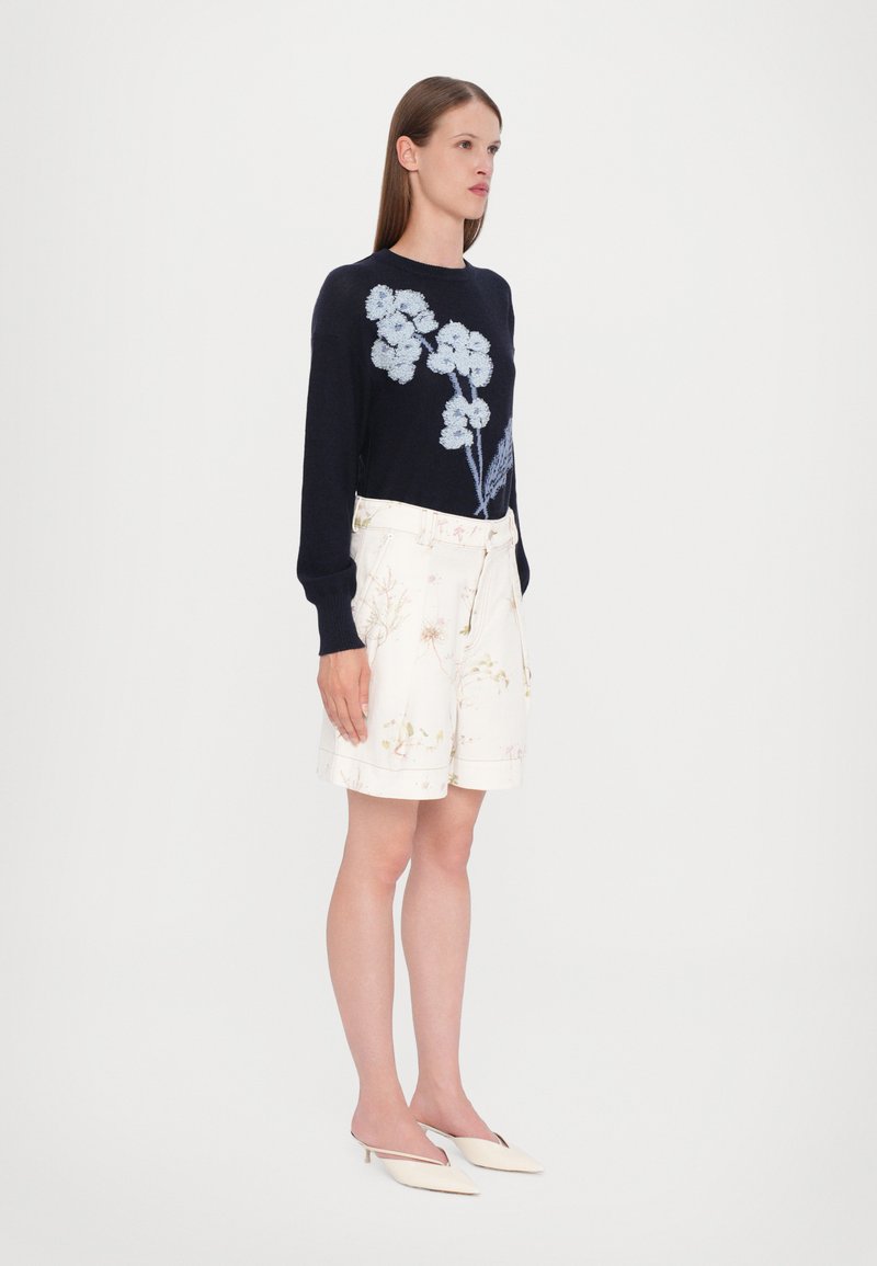 Sweater marine avec broderies florales bleues, associé à un short blanc cassé avec des motifs floraux subtils et une taille ceinturée. Talons aiguilles blancs.