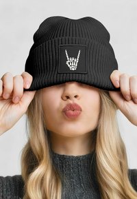 Black knitted beanie with ribbed texture and a square patch displaying a white skeleton hand graphic. The hat is pulled down slightly.