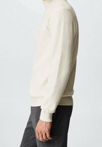 Cremfarbener, gerippter Rollkragenpullover mit figurbetonter Silhouette, aus strukturiertem Stoff und mit langen Ärmeln, kombiniert mit dunklen Hosen.