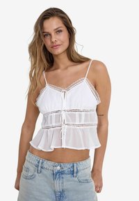 White peplum top with lace trim, adjustable spaghetti straps, and a tie-front closure, featuring a sheer lower layer and textured fabric.