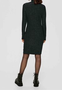 Femme portant une robe noire ajustée à manches longues et longueur genou, avec des collants noirs transparents et des bottines noires à lacets, debout de dos à la caméra.
