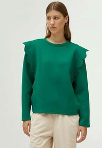 Sudadera verde con detalles ondulados en los hombros, cuello redondo, mangas largas, hecha de una tela suave, combinada con pantalones beige texturizados.