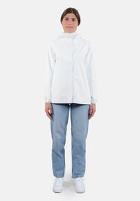 Giacca bianca leggera con cappuccio, chiusura con zip e polsini elastici, abbinata a jeans azzurri e sneakers bianche.