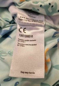 Ljusblått tyg med vattenmönster; innehåller en tvättlapp med text, CE-märke och säkerhetsmeddelande "Håll dig borta från eld."