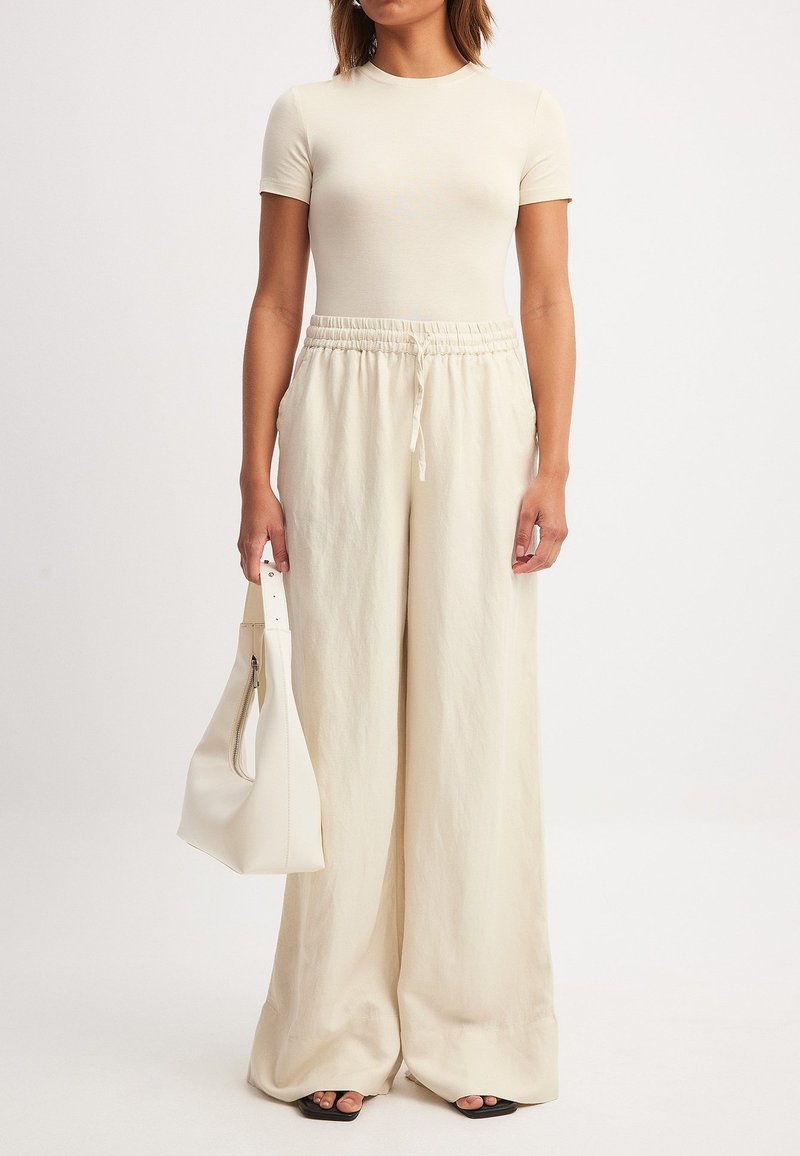 Bežs uzvalka T-krekliņš apvienots ar brīvu, krēmkrāsas platām biksēm ar elastīgu jostu. Modelis tur gludu baltu somu.