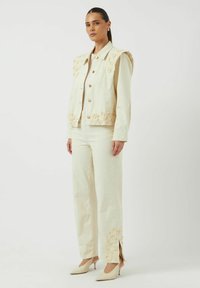 Chaqueta de mezclilla beige claro bordada con detalles florales, combinada con pantalones anchos a juego que presentan aberturas laterales y acentos decorativos.