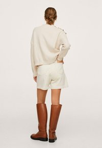 Pull beige clair en maille avec texture côtelée, associé à un short blanc et des bottes hautes en cuir marron, avec une finition lisse et des talons blocs.