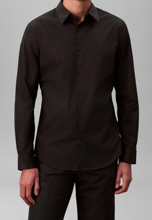 Man wearing a fitted long-sleeve black button-up shirt and black pants against a plain gray background.