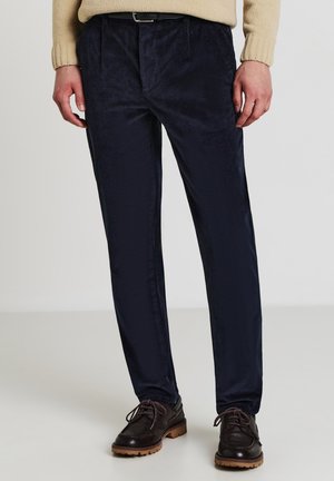 Homme portant un pantalon en velours côtelé bleu marine, un pull beige, des chaussures en cuir marron à lacets, se tenant sur un sol et un mur clairs.