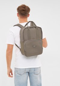 Zaino grigio con doppi manici superiori, spallacci imbottiti, tasca frontale con zip e superficie in tessuto liscio. Design minimalista con dettaglio del logo.