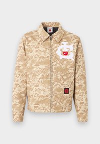 Smėlio spalvos užtraukiama striukė su šviesiai rudos kamufliažo raštu, baltu abstrakčiu piešiniu su raudonu obuoliu ant krūtinės ir mažu juodos-raudonos spalvų pleistru netoli apatinės kraštinės.