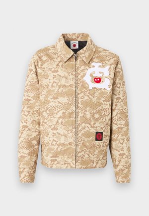 Chaqueta beige con cremallera, patrón de camuflaje marrón claro, diseño abstracto blanco con una manzana roja en el pecho y un pequeño parche negro y rojo cerca del dobladillo.