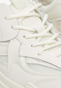 Witte synthetische sneakers hebben gaaspanelen en textuurdetails, met platte veters en zichtbare stiksels.