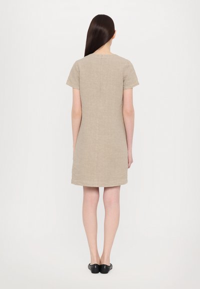 Beige linnen jurk met korte mouwen, rechte snit en rits aan de achterkant. Vlotte textuur en minimalistisch ontwerp. Zwarte schoenen zichtbaar.
