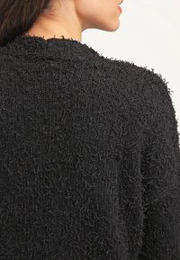 Maglione nero strutturato con un aspetto peloso e a strati. Presenta un colletto a coste e un design con schiena aperta, evidenziando il suo tessuto unico.