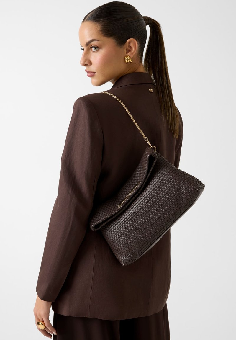 Femme avec queue de cheval foncée porte un blazer marron et des boucles d'oreilles en or, tenant un grand sac tressé marron foncé avec une bandoulière chaîne dorée sur l’épaule.