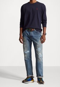 Mann in navybluse mit langen Ärmeln, verwaschenen blauen Jeans mit Flicken, gestreiftem Gürtel, bunten Sneakers und Armbanduhr, stehend vor weißem Hintergrund.